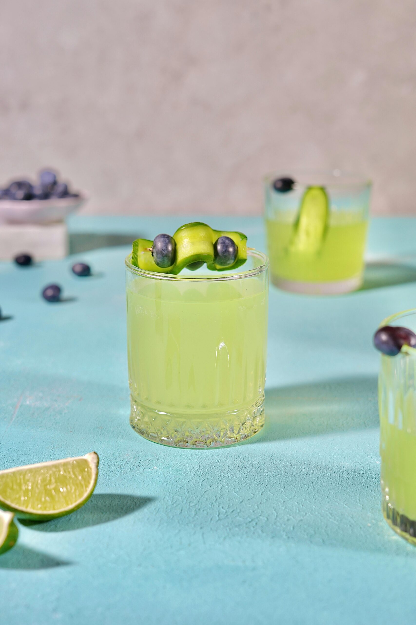Vibrant cucumber lime cocktail garnished with blueberries on textured surface.
